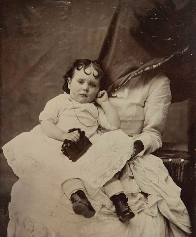 Victorian era photograph of a young girl with a calm expression in a dress. Her mother is holding her, but the mother's face is covered in a black cloth. The image is worn and yellowed from age.